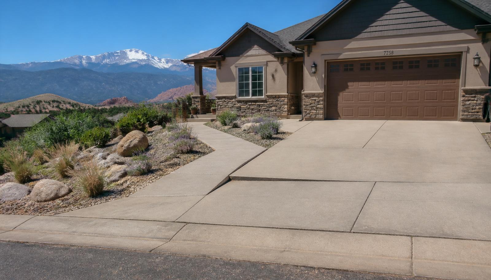 A Albuquerque residential home with a concrete driveway, front sidewalk, and side patio showing subtle signs of concrete settlement, xeriscape landscaping and the Sandias with the Rio Grande valley foothills in the background